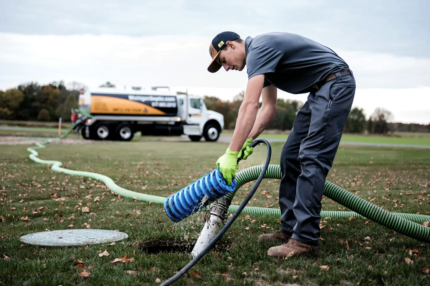 A Michigan Septic technician in work gloves handles a vacuum hose beside an open septic tank in a lawn. A pump truck lettered MichiganSeptic.com is parked behind him.