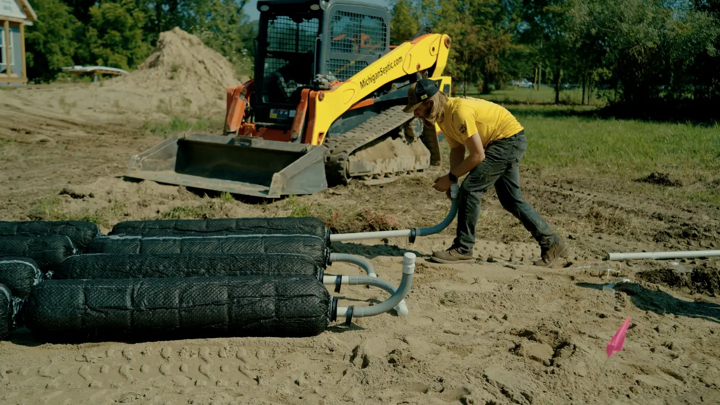 A crew member in a yellow shirt connects pipe to a row of black mesh-wrapped drainfield bundles laid out on sand. A skid steer lettered MichiganSeptic.com sits behind him.