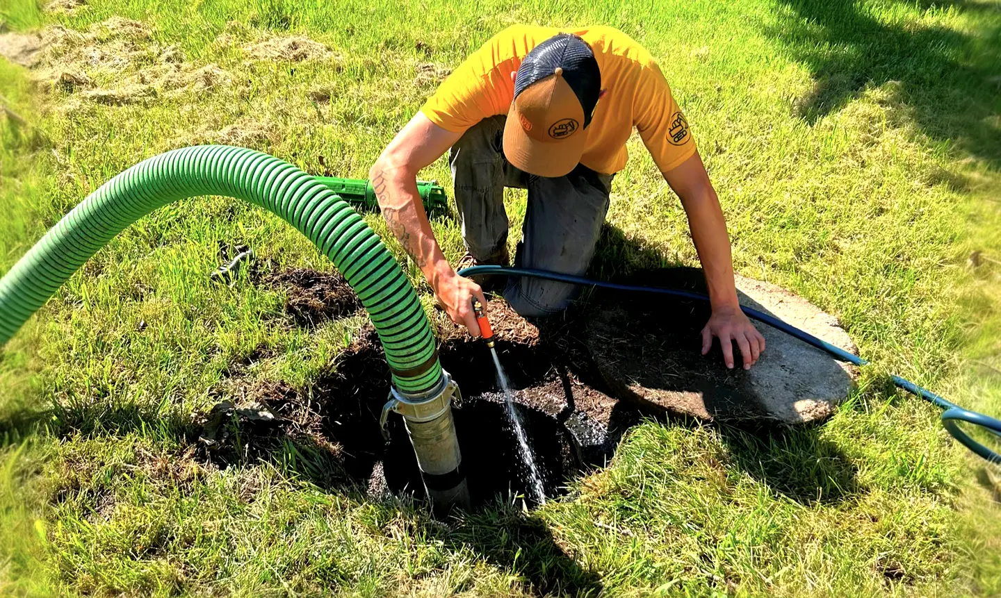 A technician in a yellow Michigan Septic shirt kneels at an open septic tank and rinses it with a hose while a green vacuum hose runs down into the tank.