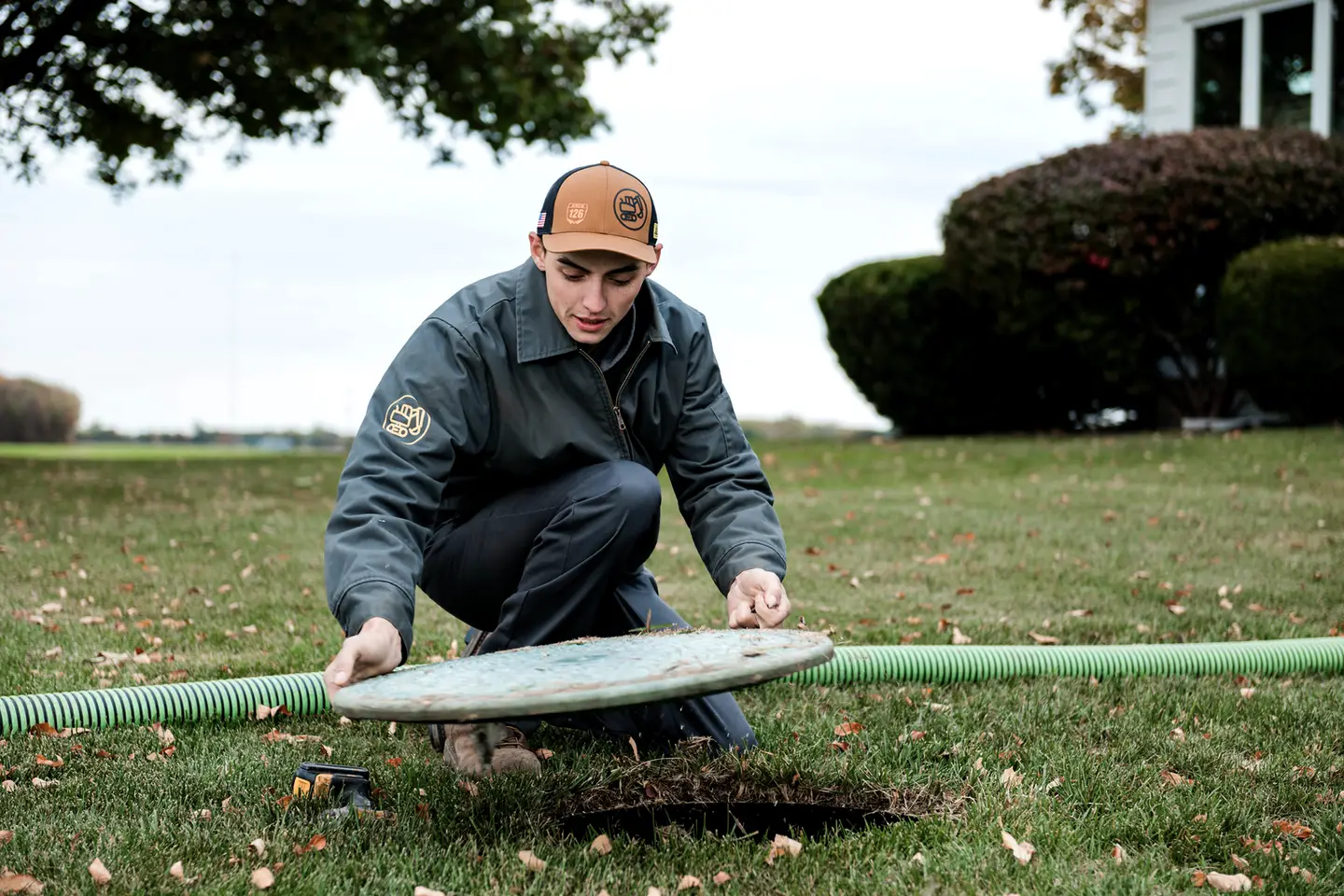 A technician crouches on a lawn and lifts the round lid off a septic tank, with a green vacuum hose lying behind him.